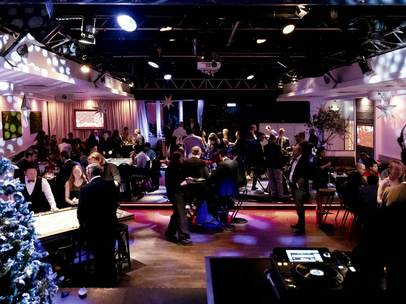 Gäste einer Firmenfeier an Spieltischen und Stehtischen, der Saal in blaues und magentafarbenes Licht getaucht